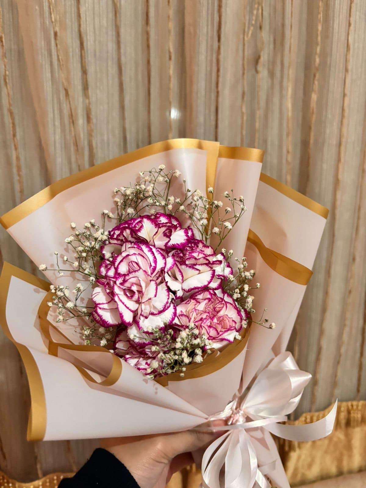 A hand holding a wrapped bouquet of magenta-edged carnations and baby's breath.